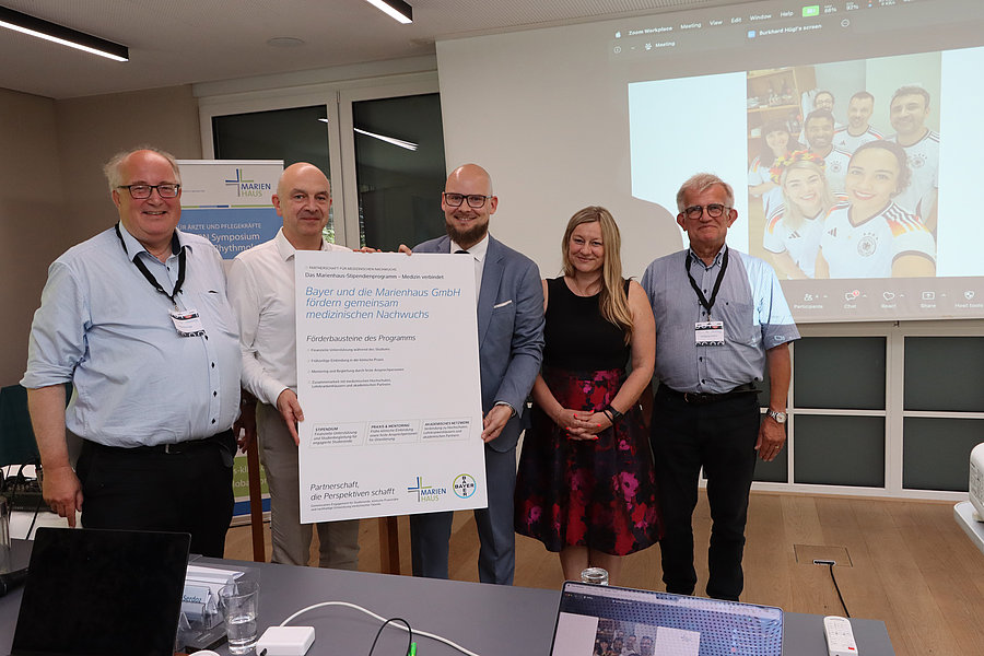 Fünf Personen stehen in einem Büro vor einem großen Plakat mit Text und lächeln in die Kamera