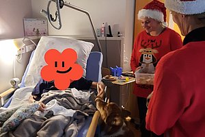 Krankenhauszimmer mit Patient im Bett, zwei Personen in Weihnachtsmützen und ein Hund am Bettende