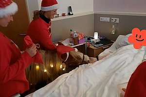 Personen in Weihnachtsmützen stehen um ein mit weißer Decke bedecktes Bett in einem Zimmer mit medizinischer Ausstattung