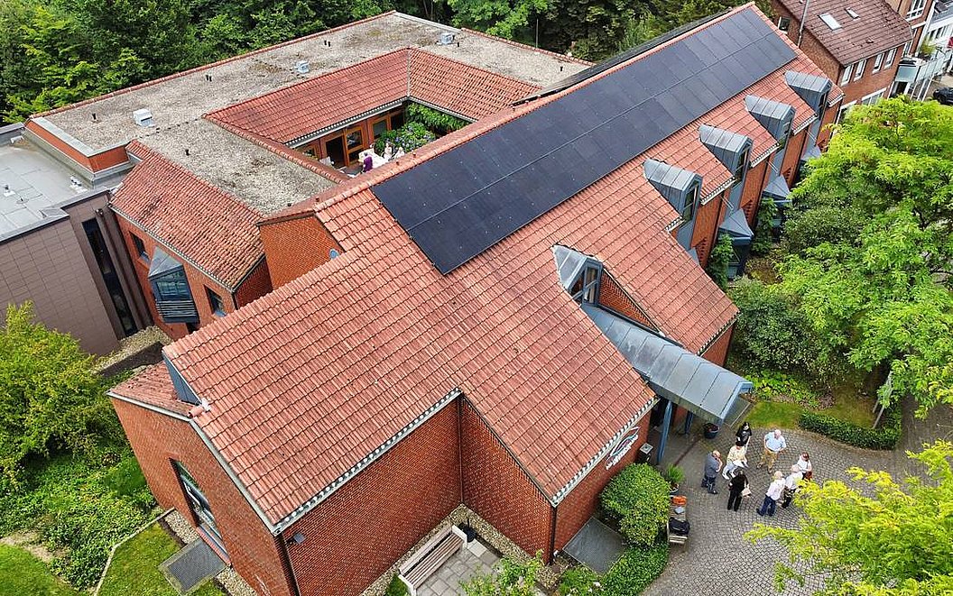 Luftaufnahme eines roten Backsteingebäudes mit Solarpaneelen auf dem Dach und mehreren Personen vor dem Eingang.