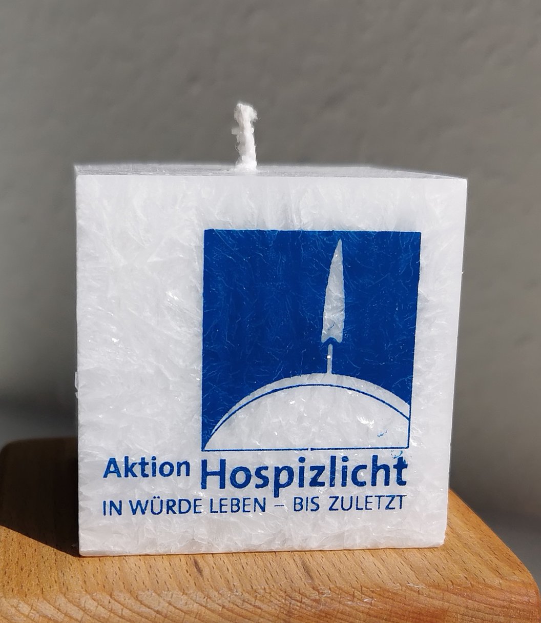 Weiße quadratische Kerze mit blauem Logo einer Flamme auf einer Halbkugel und dem Text 'Aktion Hospizlicht IN WÜRDE LEBEN – BIS ZULETZT'