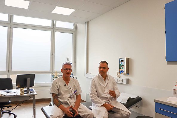 Zwei Ärzte in weißen Kitteln sitzen in einem hellen Untersuchungsraum mit großen Fenstern und medizinischer Ausstattung