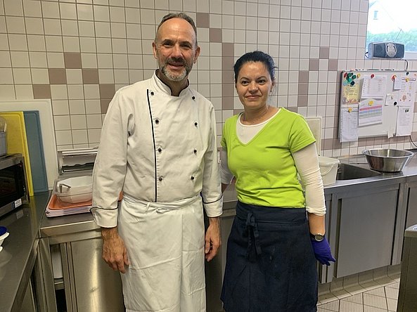 Zwei Personen in einer professionellen Küche, ein Mann in weißer Kochjacke und eine Frau in grünem T-Shirt und schwarzer Schürze, stehen nebeneinander