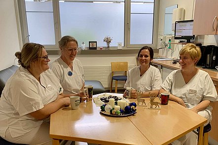 Vier Pflegekräfte in weißen Uniformen sitzen an einem Tisch in einem Pausenraum und trinken Kaffee