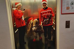 Zwei Personen in einem Aufzug, beide tragen rote Pullover, eine Person hält ein Smartphone, ein Hund mit Geschirr steht zwischen ihnen.