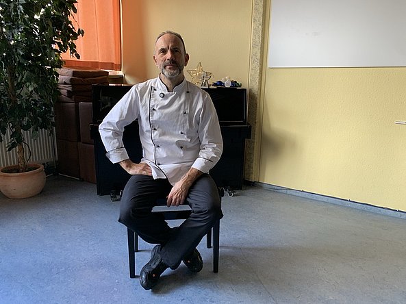 Mann in Kochjacke sitzt auf Stuhl in hellem Raum mit Klavier und Pflanze im Hintergrund