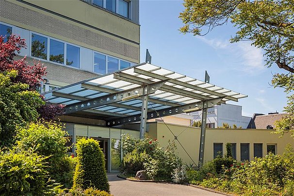 Eingangsbereich eines modernen Gebäudes mit Glasvordach, umgeben von grünen Büschen und Bäumen.