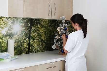 Frau in weißer Kleidung schmückt kleinen silbernen Weihnachtsbaum mit blauen und silbernen Kugeln in einem Raum mit Waldtapete