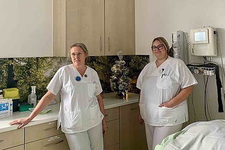 Zwei medizinische Fachkräfte in weißer Arbeitskleidung stehen in einem Behandlungsraum mit Schränken und medizinischen Geräten