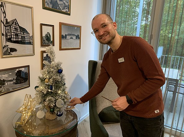 Mann mit Brille und braunem Pullover dekoriert kleinen weißen Weihnachtsbaum mit blauen und weißen Kugeln in einem Raum mit Bildern an der Wand