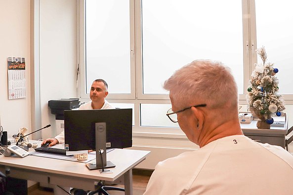 Zwei Männer in einem Büro, einer sitzt am Schreibtisch vor einem Computer, der andere sitzt mit dem Rücken zur Kamera