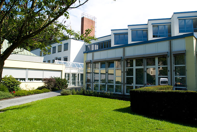 Gebäudekomplex mit Glasfassade und weißer Außenwand, umgeben von grünem Rasen und Büschen unter blauem Himmel