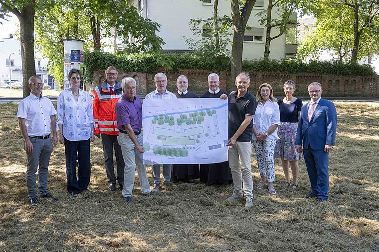 Elf Personen stehen im Freien auf einer Wiese unter Bäumen und halten gemeinsam einen großen Plan mit Gebäuden und Bäumen