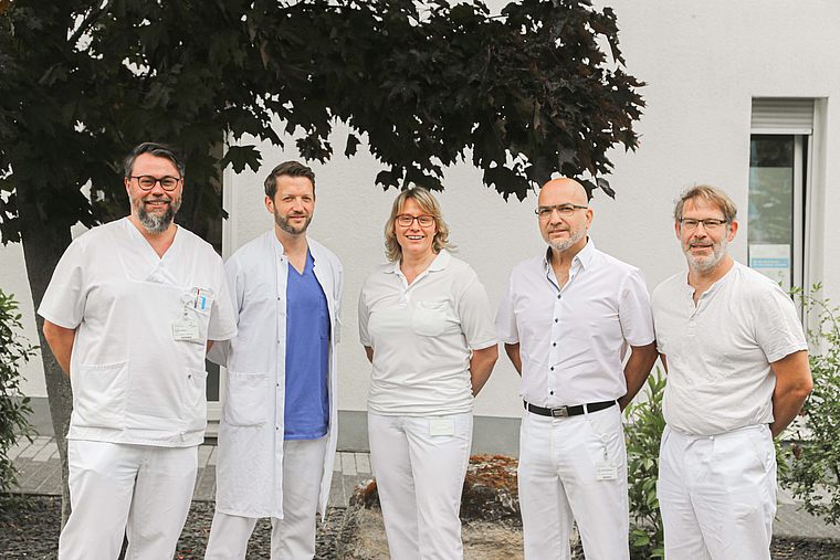 Fünf Mitarbeitende der Praxis Orthopädie Mainz in weißer medizinischer Kleidung vor einem Baum und Gebäude