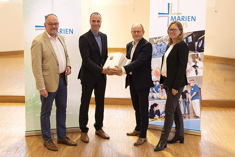 Vier Personen in Geschäftskleidung stehen vor zwei Bannern mit dem Schriftzug 'Marien' und schütteln sich die Hände, eine Person hält eine Urkunde