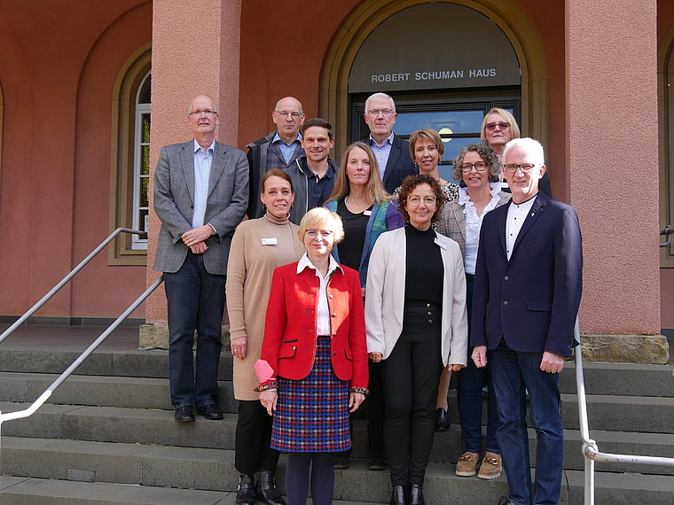 Elf Personen stehen auf Treppen vor dem Gebäude mit der Aufschrift 'Robert Schuman Haus', formell gekleidet, Gruppenfoto bei Tageslicht