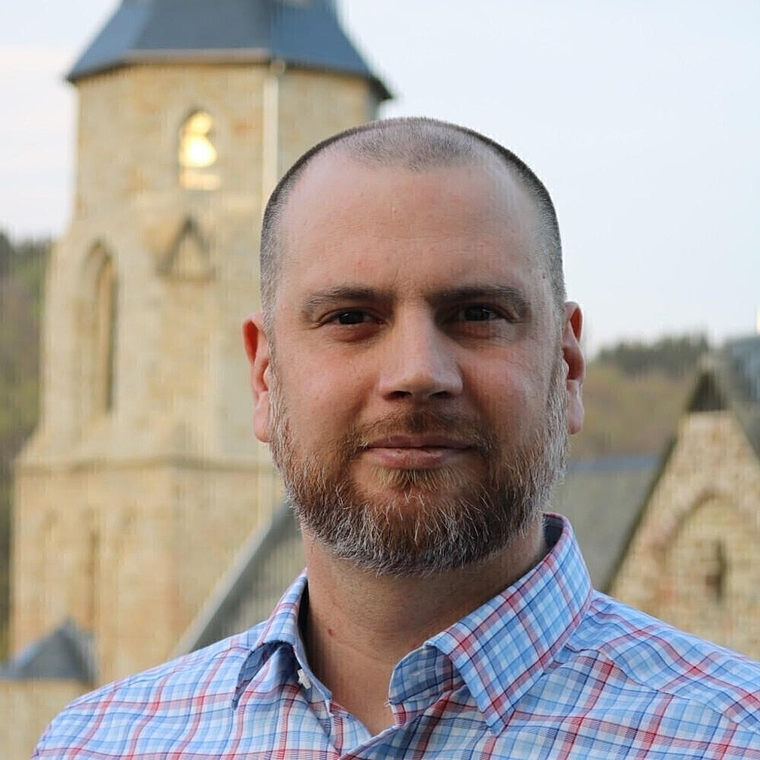 Mann mit kurzem Bart und kariertem Hemd vor einer historischen Kirche mit Turm