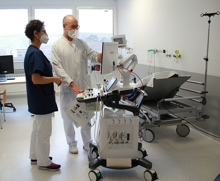 Zwei medizinische Fachkräfte mit Masken bedienen ein mobiles Ultraschallgerät in einem Krankenhauszimmer mit leerem Patientenbett