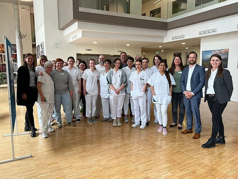 Gruppenfoto von mehreren Personen, darunter medizinisches Personal in weißer Arbeitskleidung und weitere Personen in Business-Kleidung, in einem modernen Innenraum mit Holzfußboden