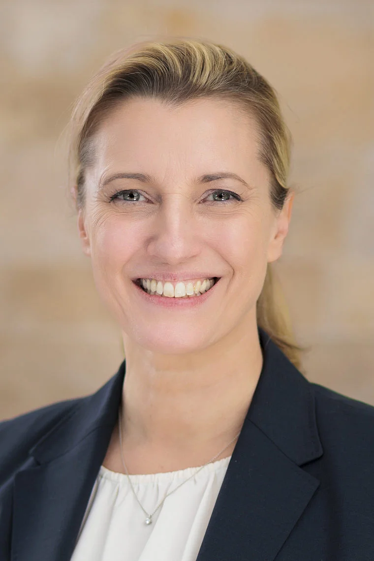 Porträt von Sandra Postel, lächelnde Frau mit blonden Haaren, trägt dunklen Blazer und weiße Bluse