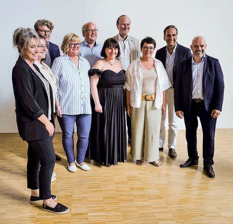 Neun Personen in formeller Kleidung bei der Eröffnung der Herzinsuffizienzeinheit, stehend auf Holzboden vor weißer Wand