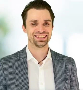 Porträt von Timo Sonntag, lächelnder Mann mit dunklem Haar, weißem Hemd und kariertem Sakko