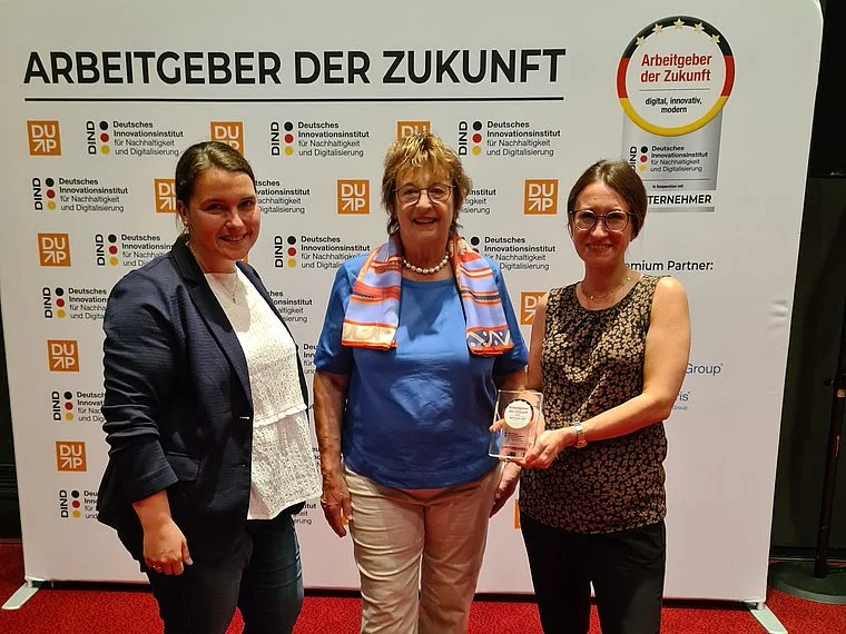 Drei Frauen stehen vor einer Wand mit dem Schriftzug 'Arbeitgeber der Zukunft' und Logos des Deutschen Instituts für Nachhaltigkeit und Digitalisierung, eine Frau hält eine Auszeichnung.