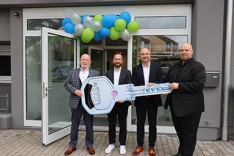 Vier Männer stehen vor einem Gebäude mit geöffneten Glastüren und halten gemeinsam einen großen Schlüssel mit der Aufschrift 'BKS'. Über der Tür sind blaue, grüne und silberne Luftballons angebracht.