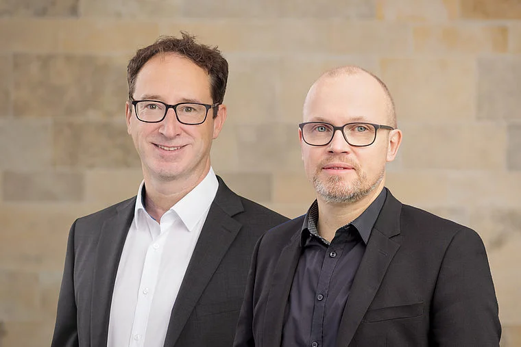 Nietzel und Link, zwei Männer in Anzügen mit Brillen, vor einer hellen Steinwand