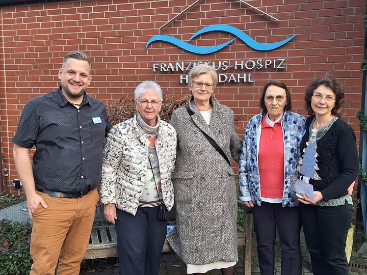 Fünf Personen stehen vor einer Backsteinwand mit dem Schriftzug 'FRANZISKUS-HOSPIZ HILDDAHL', zwei Frauen tragen gemusterte Jacken, eine Frau einen langen Mantel, ein Mann ein dunkles Hemd und eine weitere Frau einen schwarzen Pullover mit Schal.