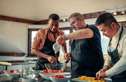 Drei Personen mit Schürzen bereiten in einer Küche gemeinsam Speisen zu, eine gießt Flüssigkeit in eine Schüssel mit Tomaten, eine schneidet Gemüse
