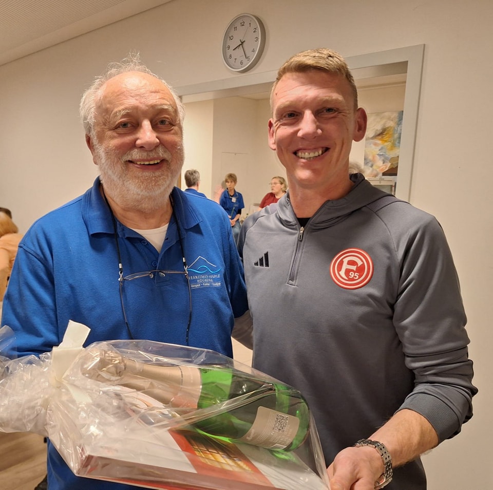 Zwei Männer stehen nebeneinander, einer trägt blaues Hemd mit Brille am Kragen, der andere graues Adidas-Sportshirt mit Fortuna-Düsseldorf-Logo, sie halten eine Geschenkverpackung mit Flasche und Buch.