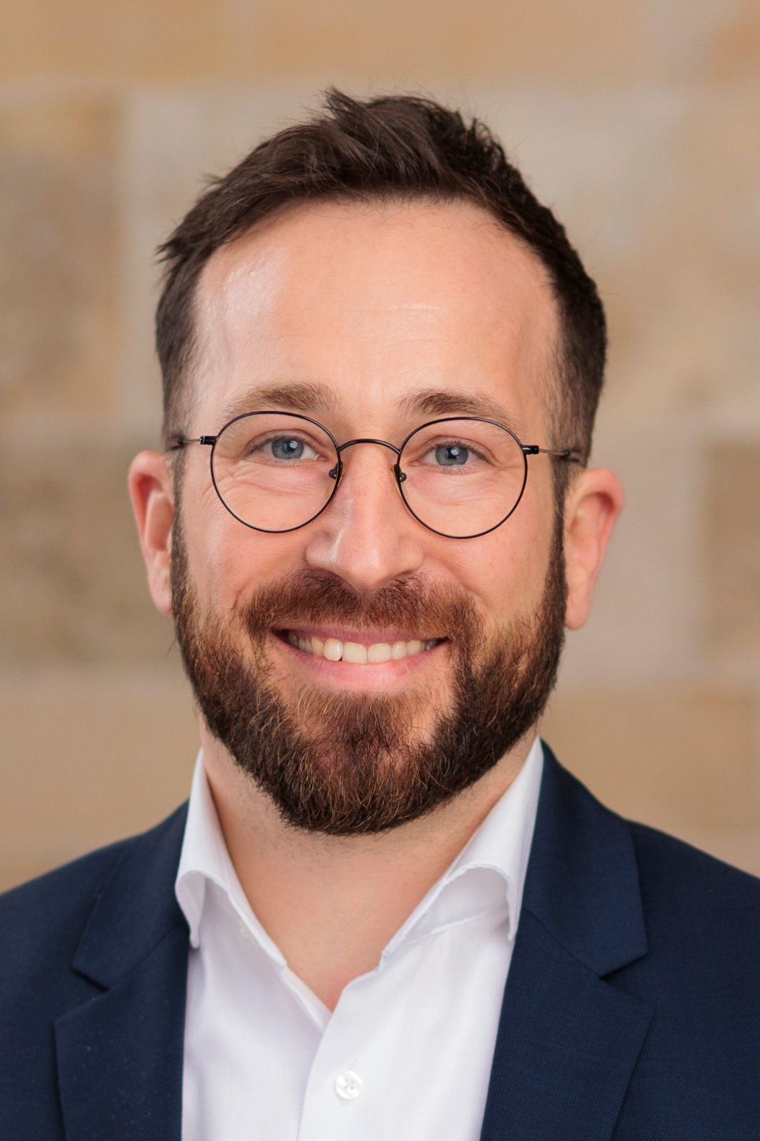 Sebastian Spottke mit Brille, kurzem braunem Haar und Bart, lächelt, trägt weißen Hemdkragen und dunklen Anzug